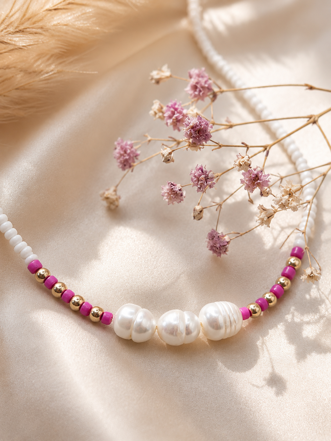 Handgemaakte witte kralenketting met drie zoetwaterparels, fuchsia accenten en goudkleurige details op een satijnen ondergrond met gedroogde bloem als decoratie