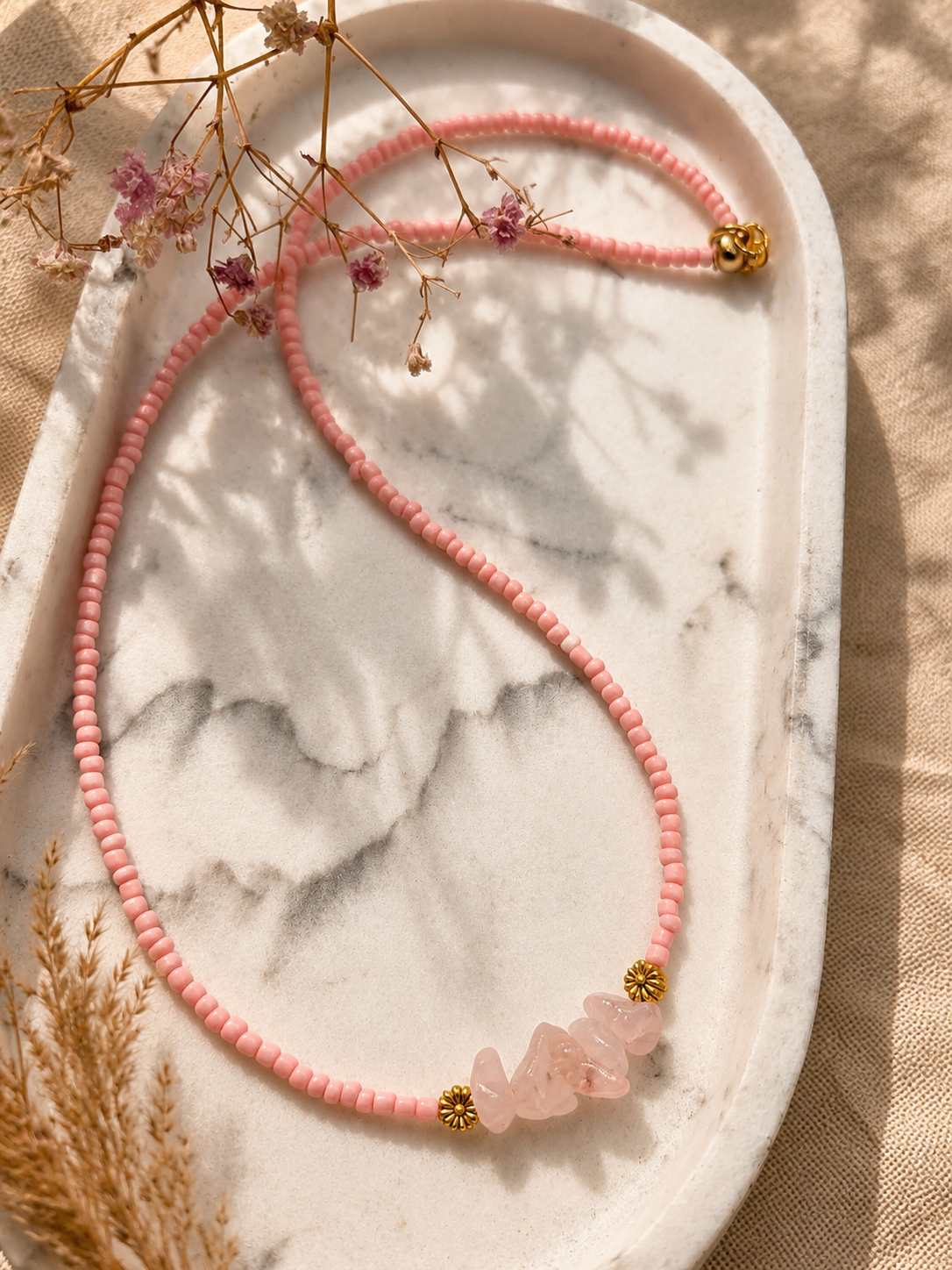 op een marmeren plaat in een zonnige sfeer ligt een lichtroze kralenketting met rozenkwarts edelstenen en goudkleurige bloemaccenten