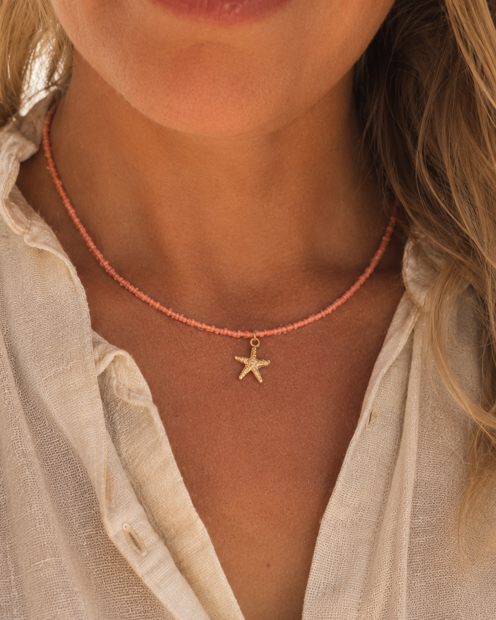 blonde vrouw met linnen blouse draagt een subtiele koraalroze kralenketting met een gouden zeesterbedeltje