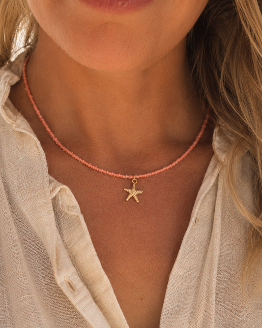 blonde vrouw met linnen blouse draagt een subtiele koraalroze kralenketting met een gouden zeesterbedeltje