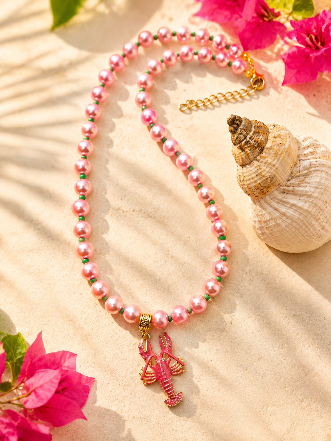 Handgemaakte roze kralenketting met goudkleurige kreeft bedel op een lichte ondergrond met bloemen en schelp.