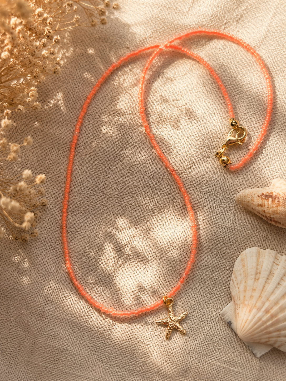 op zacht linnen doek met schelpen ligt een neon koraalroze kralenketting met een gouden zeester bedeltje