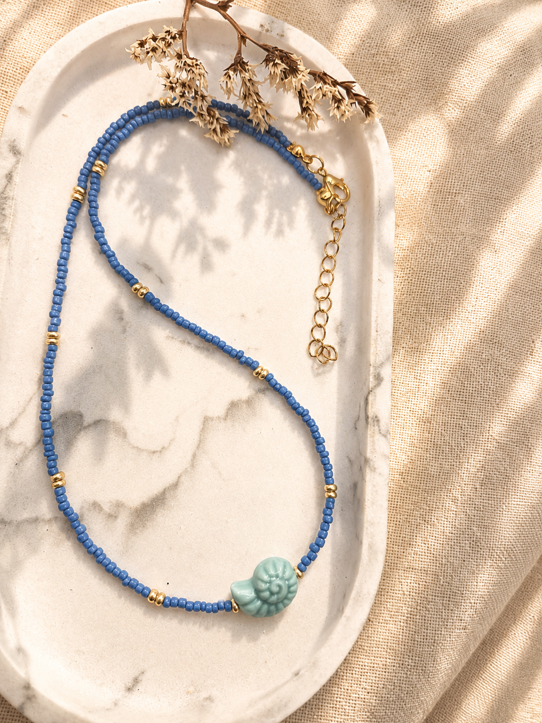 Blauwe kralenketting met goudkleurige details en een opvallende lichtblauwe schelpvormige hanger, gefotografeerd op een wit schaaltje.