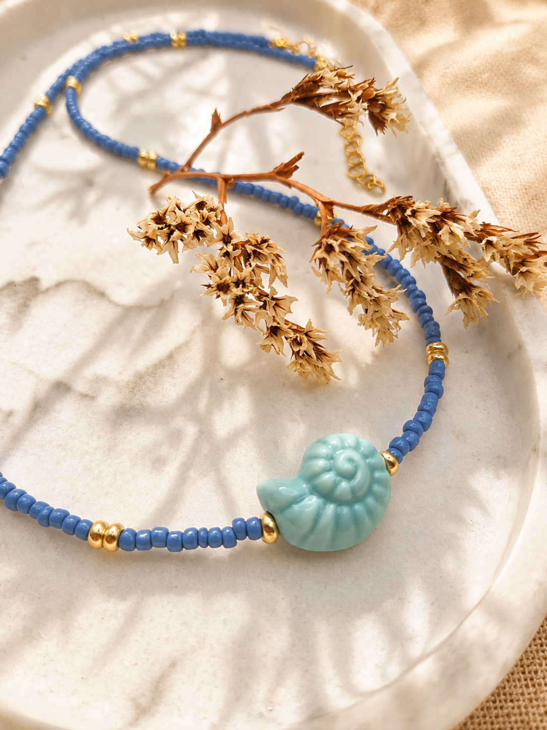 Detail van een blauwe kralenketting met goudkleurige tussenkraaltjes en een lichtblauwe schelpvormige hanger.