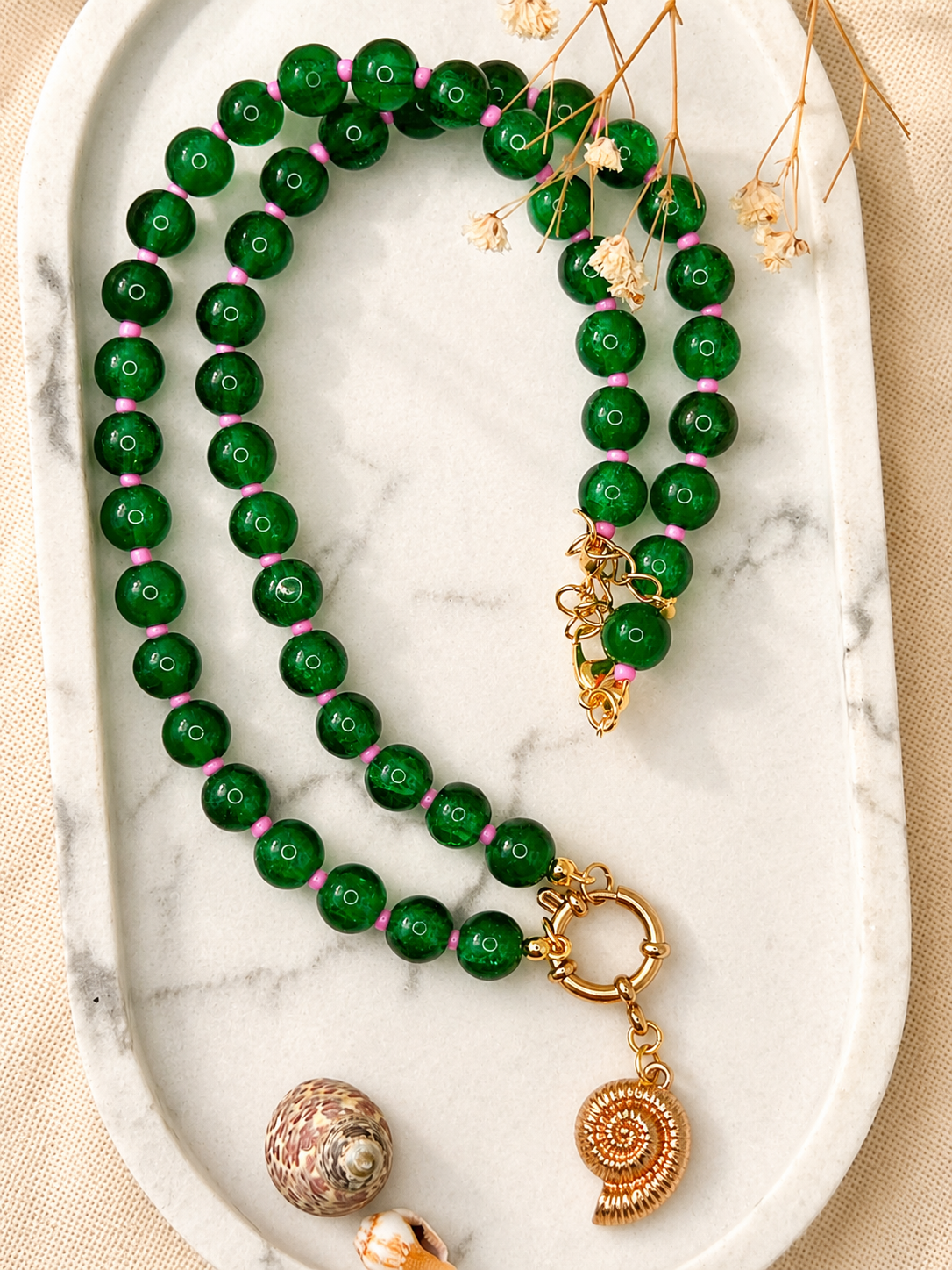 een smaragdgroene kralenketting met roze kleine tussenkraaltjes op een marmeren plaat met schelpjes met met een gouden boeislot en schelpbedel 