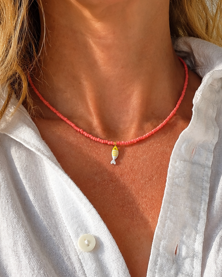een zongebruind model met blonde haren en een witte linnen blouse draagt een koraalroze ketting met een klein wit en geel gestreept visbedeltje