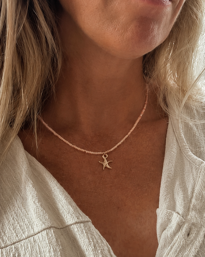 zongebruind model met blond haar draagt een koraalroze neon kralenketting met zeester bedel