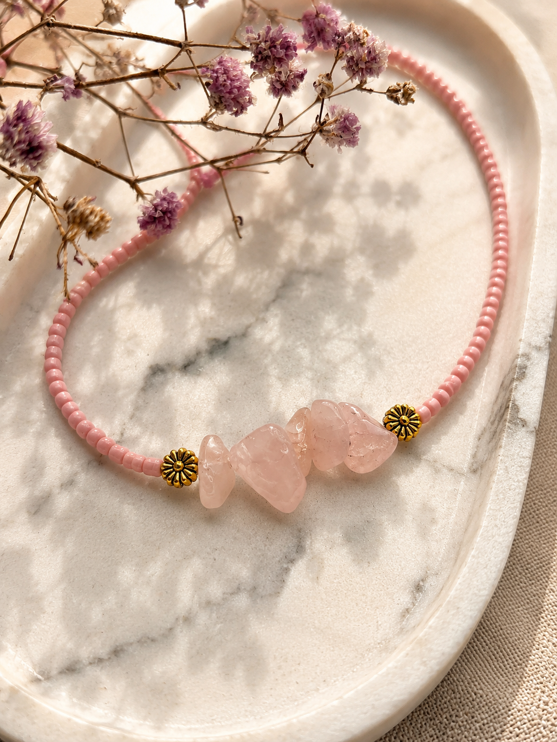 op een marmeren plaat ligt een roze kralenketting met edestenen van rozenkwarts en goudkleurige accenten met een mooie roze gedroogde bloem ernaast