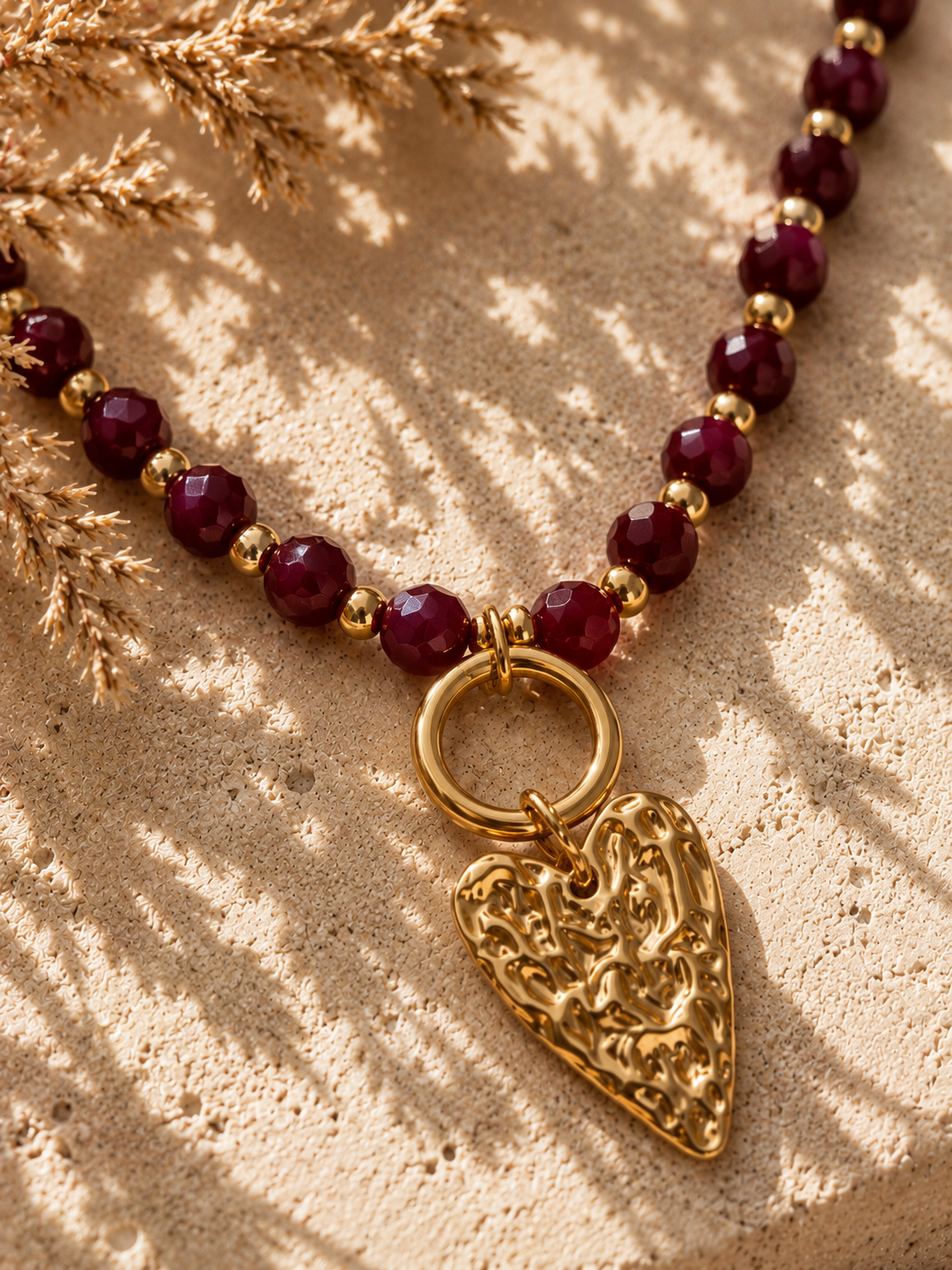 Detailfoto van een bordeauxrode kralenketting met goudkleurige hartbedel en facetgeslepen kralen.